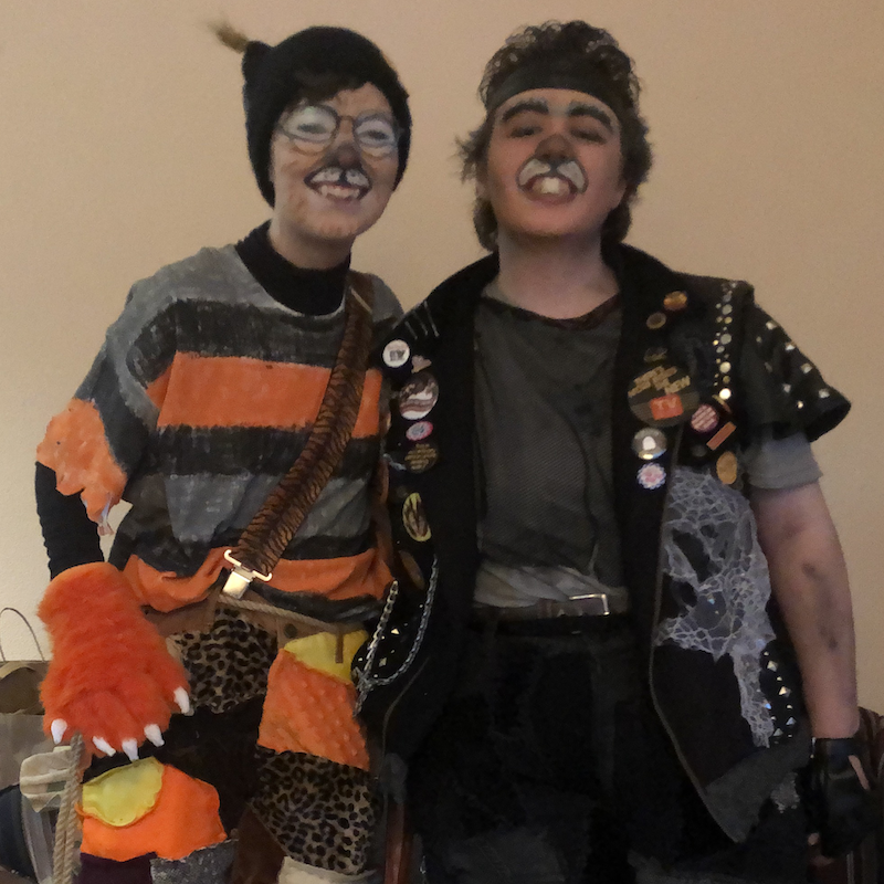 Two people, one wears a scruffy orange striped shirt, pants, and has cat paws and makeup. The other wears scruffy black pants, denim vest, and rat makeup. Link takes you to project page.